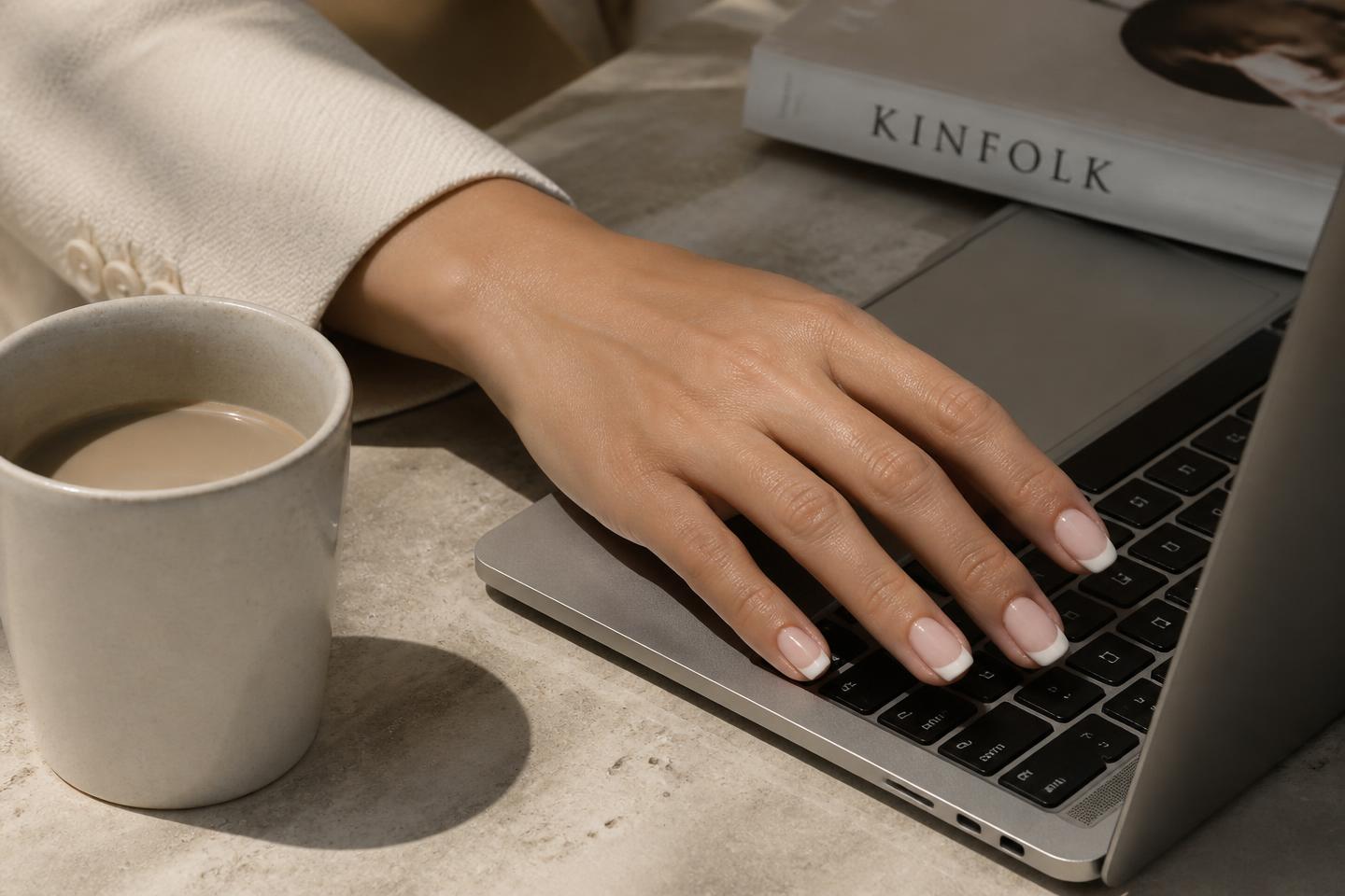 Hand with french-tip press-on nails resting on a laptop beside a coffee cup